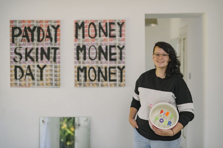Amanda Lynch standing in the far right of the image with dark shoulder-length hair, glasses, black sweatshirt with white band. Behind figure and to the left are art works, Money money money mixed media work, pay day skint day mixed media art work. Mirror propped on wall bottom left, with cement cast of feet at the bottom