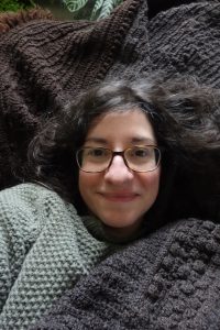 A young white woman with glasses and dark brown hair lies back on a pile of grey crocheted throws, and has more throws piled on top of her. Only her smiling face is visible.