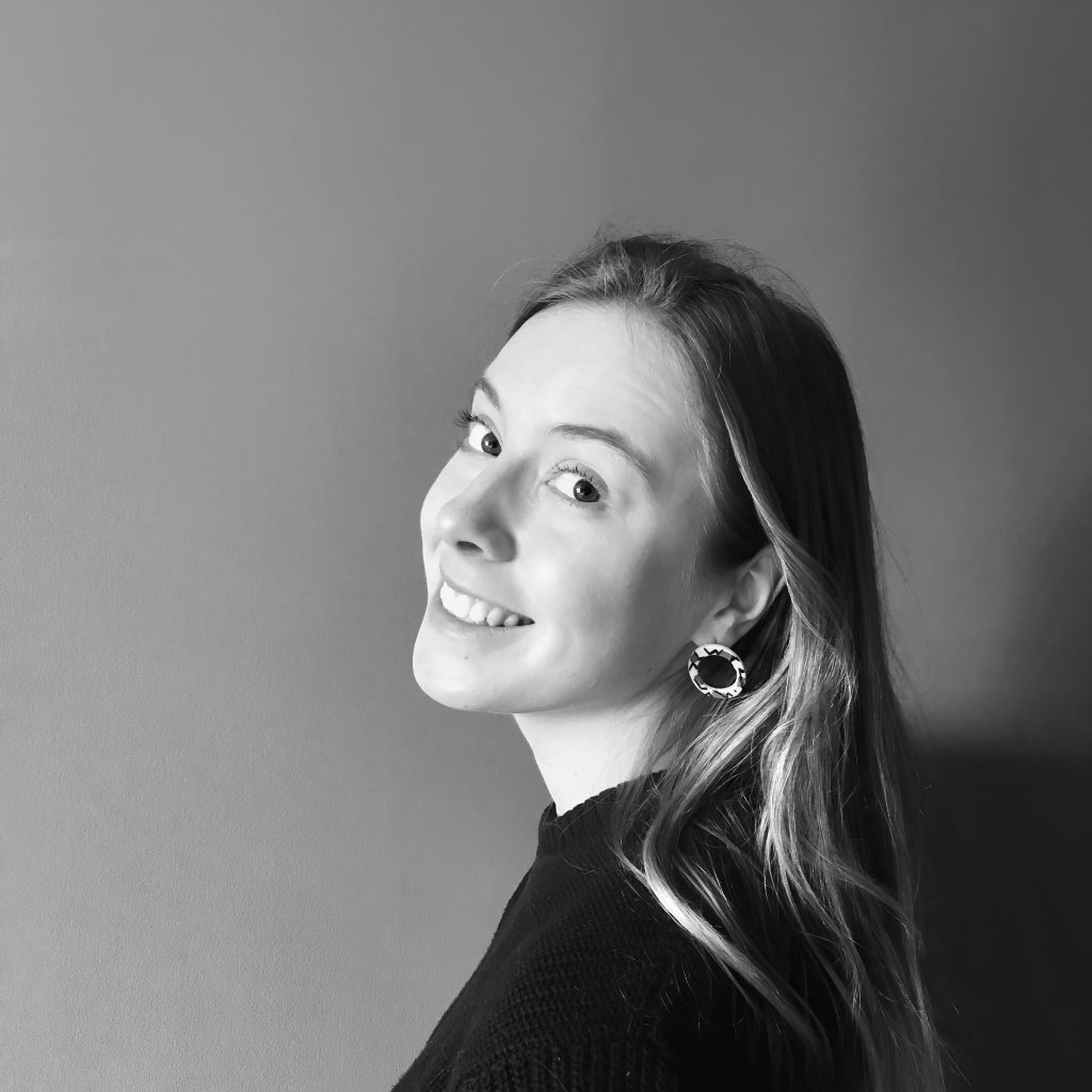 A black and white photo of Elspeth, a young white woman, looking back over her shoulder and smiling.