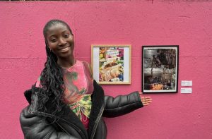 A stylishly-dressed young woman standing in front of 2 collages on a pink wall. She is smiling and gesturing