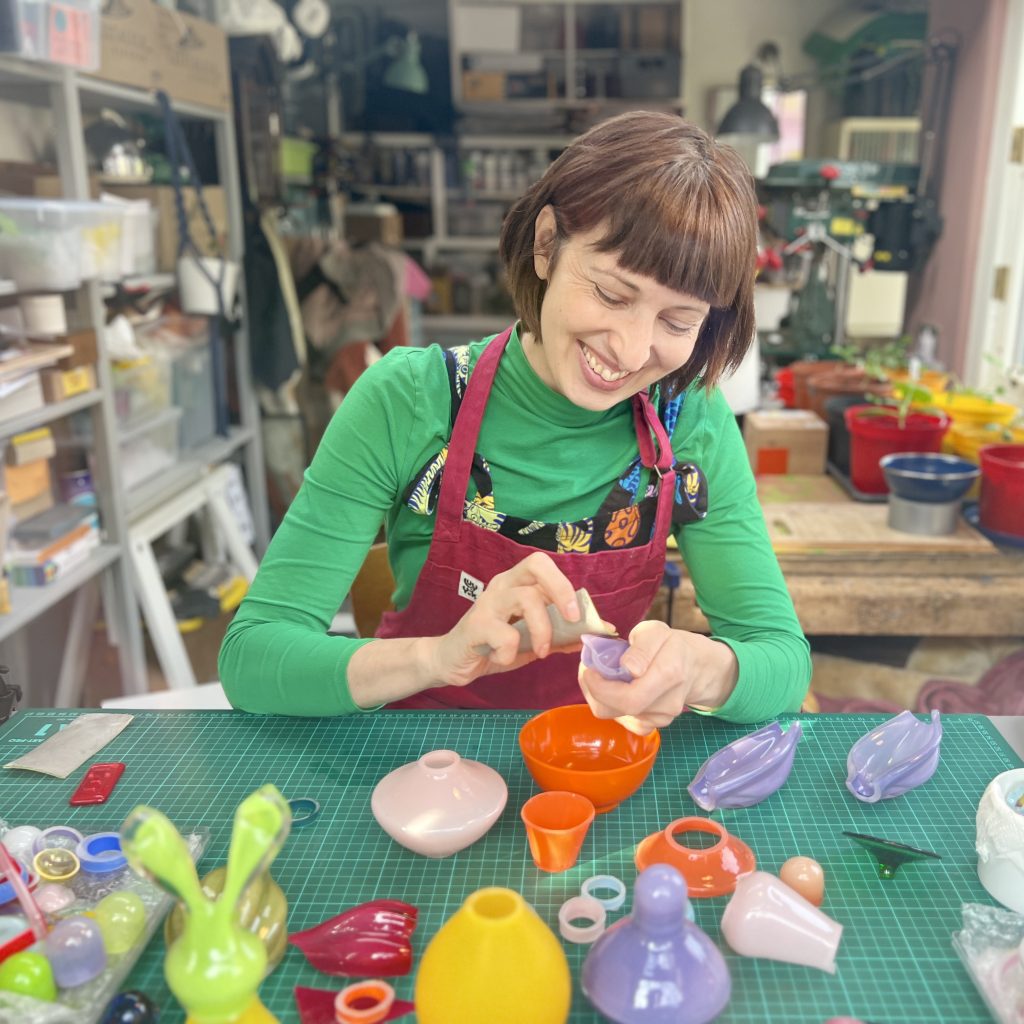 A smiley young white woman with short brown hair sits at her desk, working on vibrant glass sculptures
