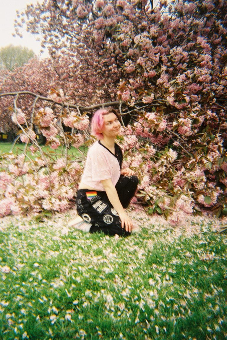 A non binary person with pink and brown hair kneeling in front of a fallen cherry blossom tree. The scene is very pink and the person is dressed in punk clothing