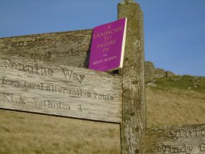 A magenta book nestles against a tri-directional wooden Pennine Way sign, with grassland, crags and blue sky in the background