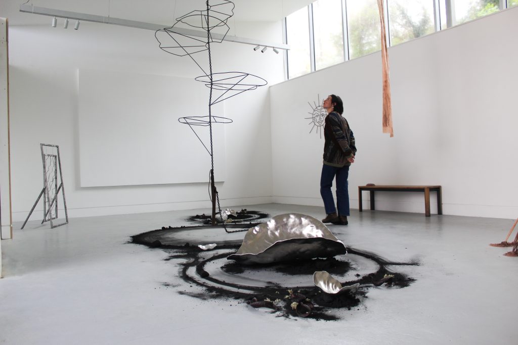 A collection of metalwork sculptures, surrounded by black sand trailing across the floor, with a central hanging piece of thin rod around which a person walks.