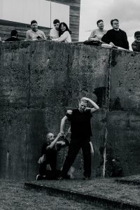 Black and white photo of a performance, taking place in what looks like a concrete pit. One man is kneeling, clinging to another (standing) man, who also seems to have another performer tucked behind them