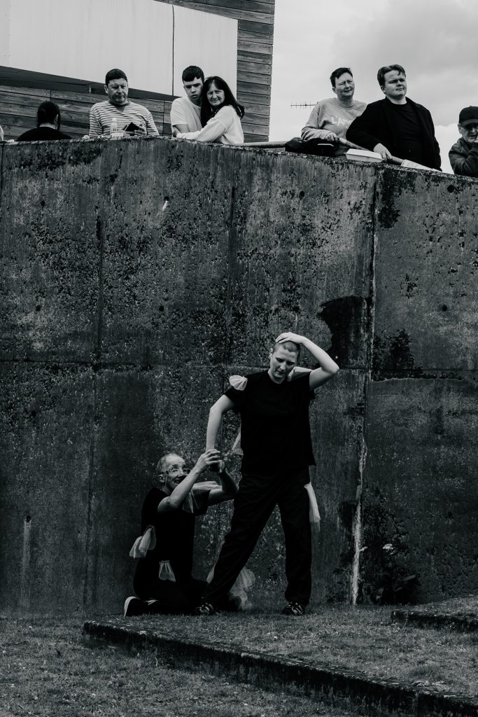Black and white photo of a performance, taking place in what looks like a concrete pit. One man is kneeling, clinging to another (standing) man, who also seems to have another performer tucked behind them