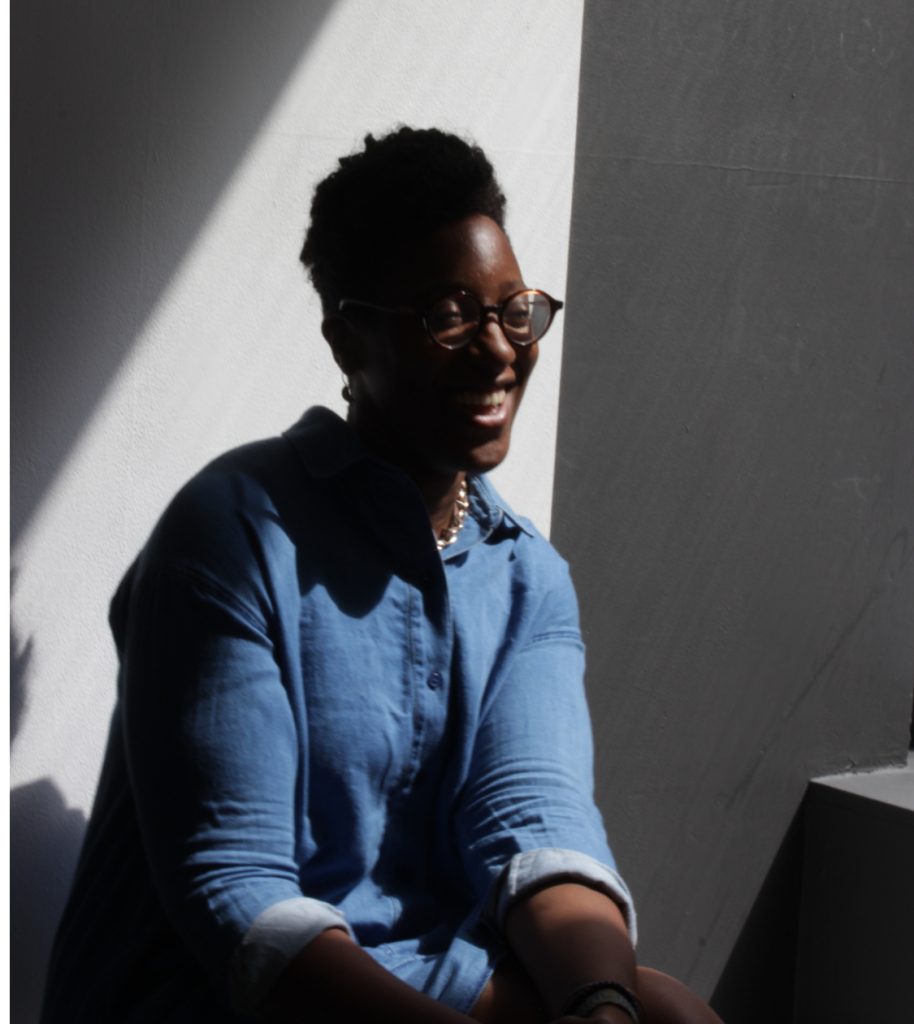A Black woman with curly short cropped hair, wearing a blue denim dress smiling against a sun lit white background with a shadow cast in the background