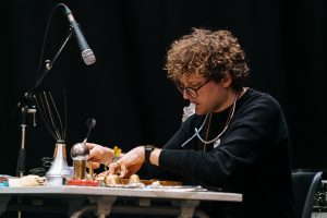 Ali Robertson, a white man with curly brown hair and glasses, sits at a table playing an array of found objects as musical instruments, including a head massager, a trumpet mute, a metal ball, some springs, a bell, a miniature toy kettle, a hand whisk, and more. A microphone is positioned above him, pointed at the table, while he holds an empty pill blister packet between his lips.