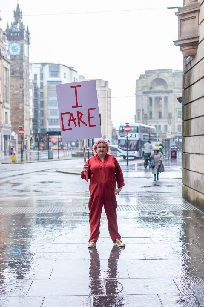 A short woman with curly hair, wearing a red boilersuit holds a placard that says I CARE