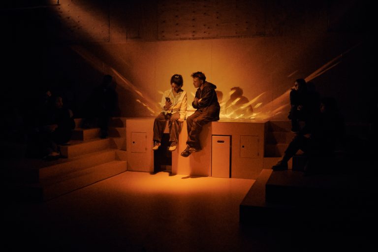 A piece of installation theatre, the space is in a bright orange box. Two actors sit on an orange podium surrounded by fractured light and audience surround in the shadows.
