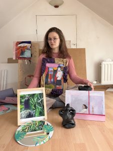 The image shows a young woman with glasses, seated on a wooden floor in an attic-like room, surrounded by her artwork. She holds a framed piece featuring a colourful painting of a nude figure, while in front of her is a framed bird painting, a circular artwork with tropical leaves, and a black abstract sculpture. Additional artwork, including a vibrant townscape painting, leans against the wall behind her.