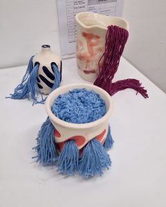Three ceramic pieces of different shapes and heights have wool pouring out of spouts or holes cut into the walls of the vessels. The pieces are a mixture of pinks and blues.