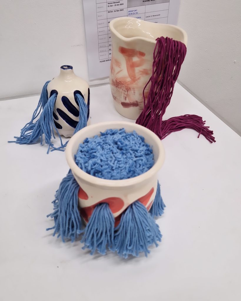 Three ceramic pieces of different shapes and heights have wool pouring out of spouts or holes cut into the walls of the vessels. The pieces are a mixture of pinks and blues.