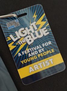A blue rectangular lanyard for Light The Blue festival 2024 with the designation of Artist. It has a yellow lightening flash across it and text that says 'For Young People, By Young People'.