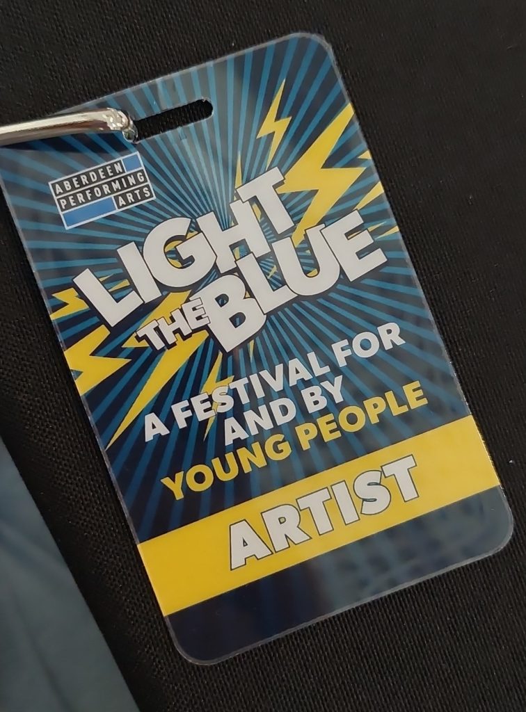 A blue rectangular lanyard for Light The Blue festival 2024 with the designation of Artist. It has a yellow lightening flash across it and text that says 'For Young People, By Young People'.