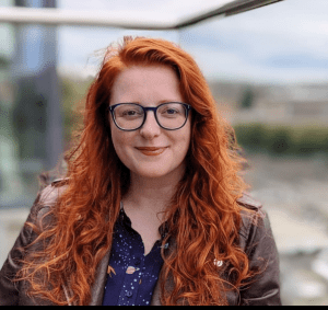 A woman with pale, white, freckled skin, and long, vibrant red, wavy hair, is smiling at the camera. She's wearing round blue glasses, a cobalt blue top with pink and yellow rockets and planets on it and a brown faux leather jacket. She has blue eyes, and is sitting on a balcony with blurred hills in the distance.