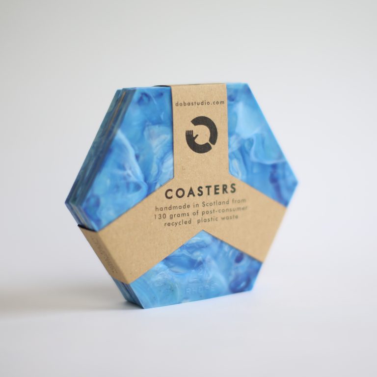 The image shows a hexagonal set of coasters made from recycled plastic waste. The coasters are vibrant blue, with swirling, marbled patterns giving them a distinctive and artistic look. They are neatly packaged with a brown cardboard sleeve, featuring clean, modern typography.