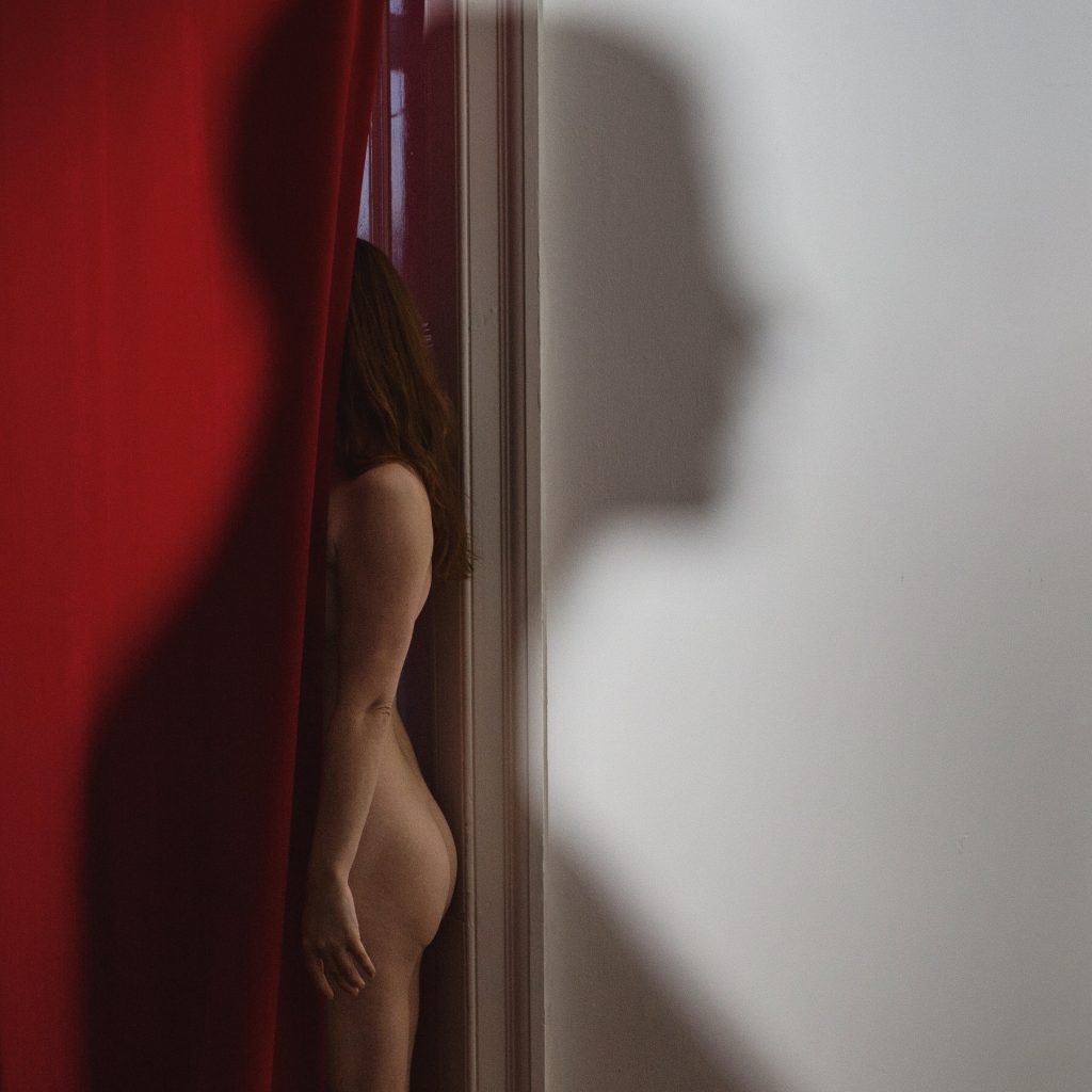 Image of a naked female form hidden behind a red velvet curtain with the shadow of a male figure across the picture.