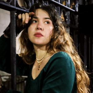 A portrait photograph of a white woman with long brown hair, wearing a dark green dress, posing between the bars of a fence. The camera angle is looking up at her and she is looking into the distance.