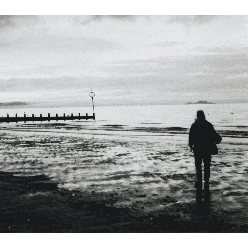 A black & white image of a beach during a cloudy day. There is a silhouette of a person in the foreground, and there’s a wooden tide measure in the background.