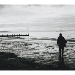 A black & white image of a beach during a cloudy day. There is a silhouette of a person in the foreground, and there’s a wooden tide measure in the background.