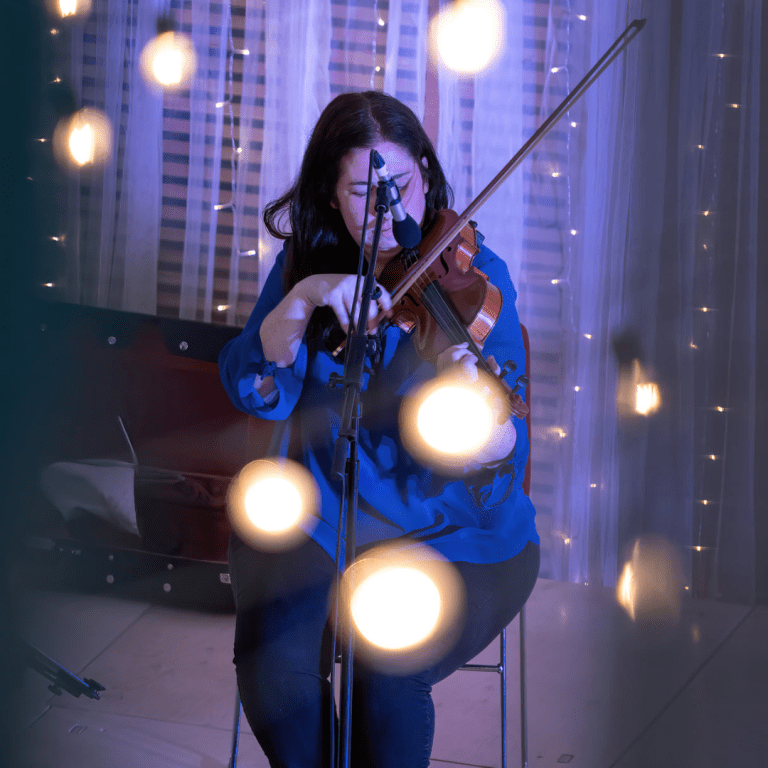 Photo of Eilidh. She has brown hair, a blue top and is playing the fiddle on stage at a concert.