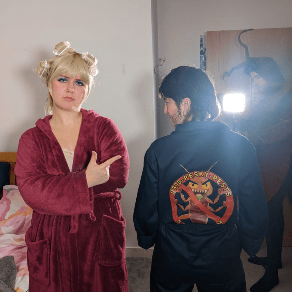 A person in a red dressing gown with curlers in their hair is facing the camera and pointing to a person standing on their right, who is wearing dark overalls and has their back to the camera. There is a graphic design on the back of their overalls.