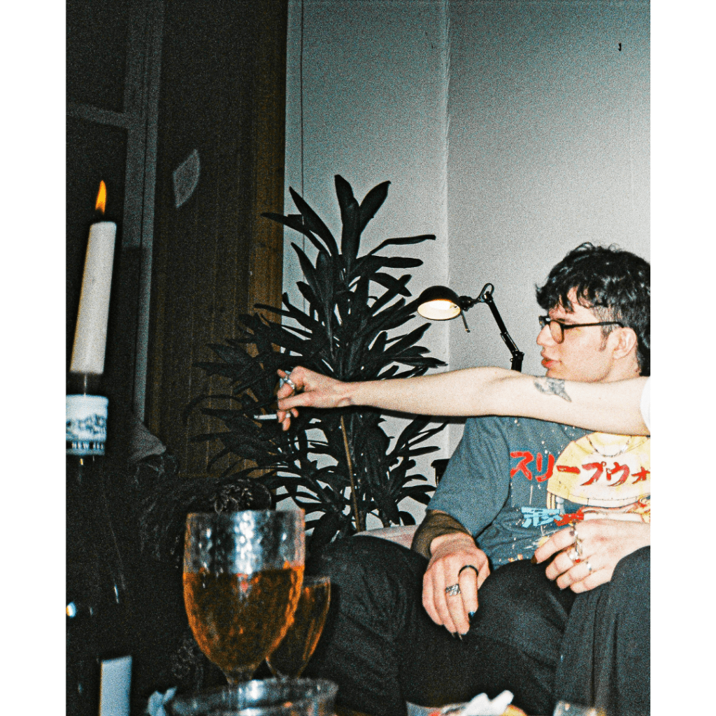A flash photo of a party, candles and glasses of wine on the table. A man sits looking to the left while another person’s arm stretches in front of him into middle frame holding a cigarette.