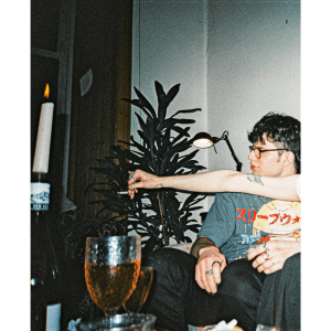 A flash photo of a party, candles and glasses of wine on the table. A man sits looking to the left while another person’s arm stretches in front of him into middle frame holding a cigarette.