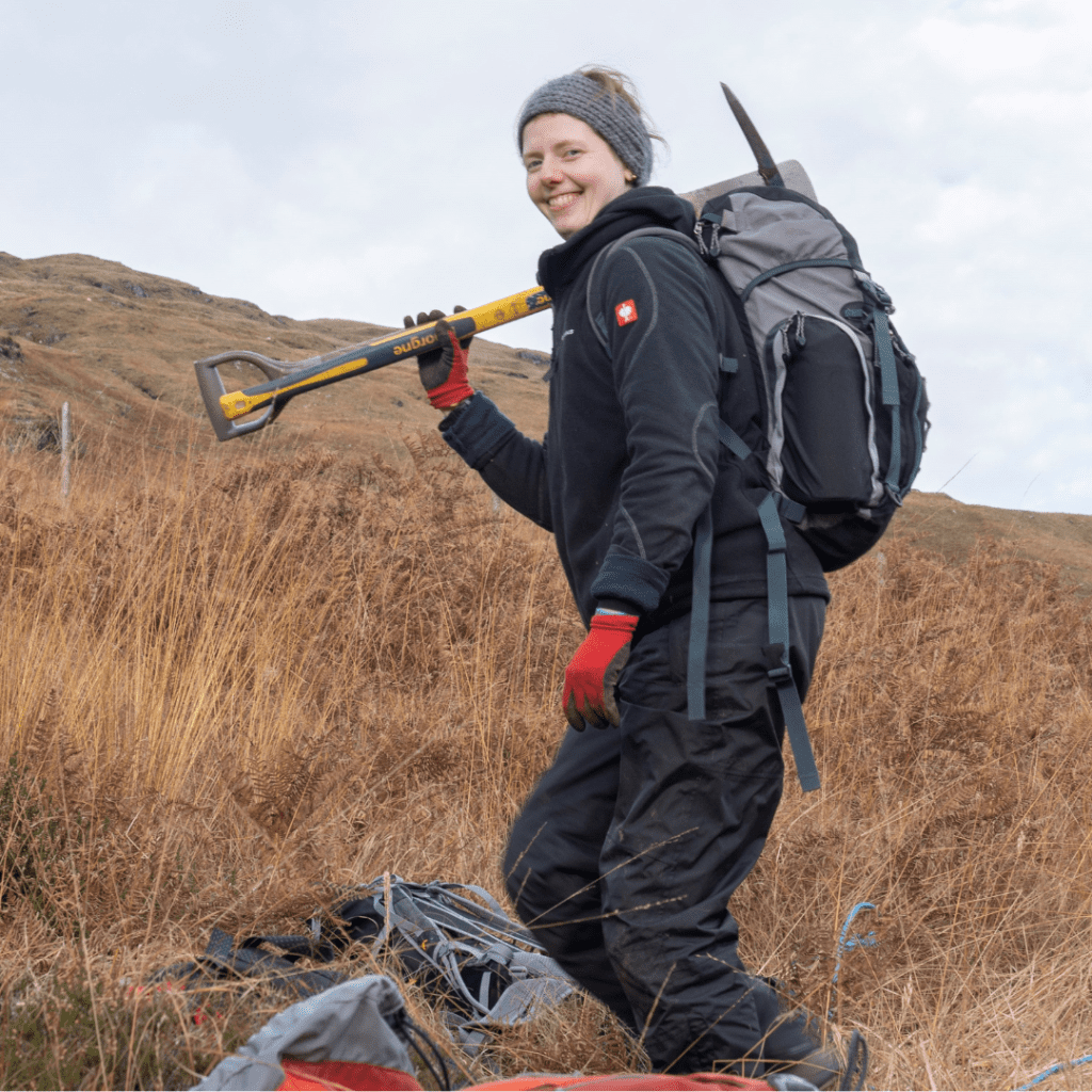 A person wearing black waterproofs and a rucksack is smiling at the camera. They are standing on a grassy hill carrying a pickaxe.