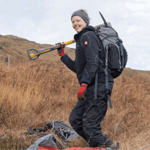 A person wearing black waterproofs and a rucksack is smiling at the camera. They are standing on a grassy hill carrying a pickaxe.