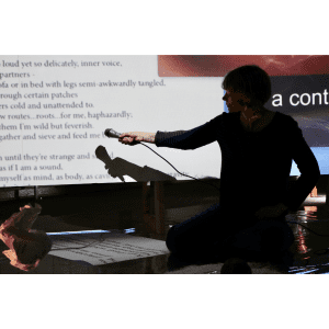 Image shows a close-up of a silhouetted person wearing dark clothing, kneeling in front of an upstanding projection screen and on floor that is covered with reflective material. Their arm is outstretched towards the screen, holding a microphone towards a projection of poetic text.