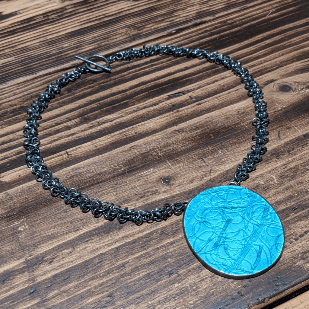 Green, enamelled, oval pendant with fireweed pressed into the surface on a dark grey multi-link chain arranged in a circle on a wooden surface