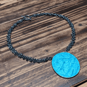 Green, enamelled, oval pendant with fireweed pressed into the surface on a dark grey multi-link chain arranged in a circle on a wooden surface