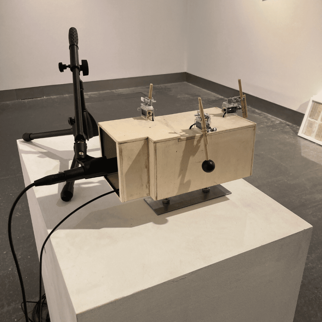A scale model of a room made of birch plywood situated on a white plinth. A microphone is positioned at the room entrance and there are small machine controlled mallets on three of the four walls.