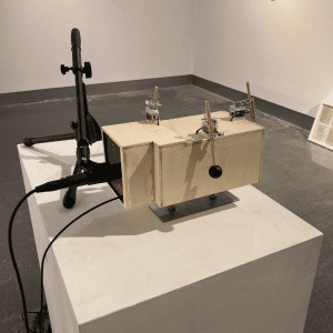 A scale model of a room made of birch plywood situated on a white plinth. A microphone is positioned at the room entrance and there are small machine controlled mallets on three of the four walls.