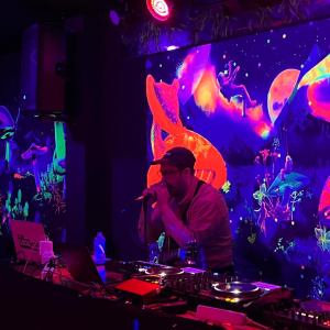 A man wearing a baseball cap speaks into a microphone. He is standing behind a DJ desk with a projection of mushrooms behind him in fluorescent colours.