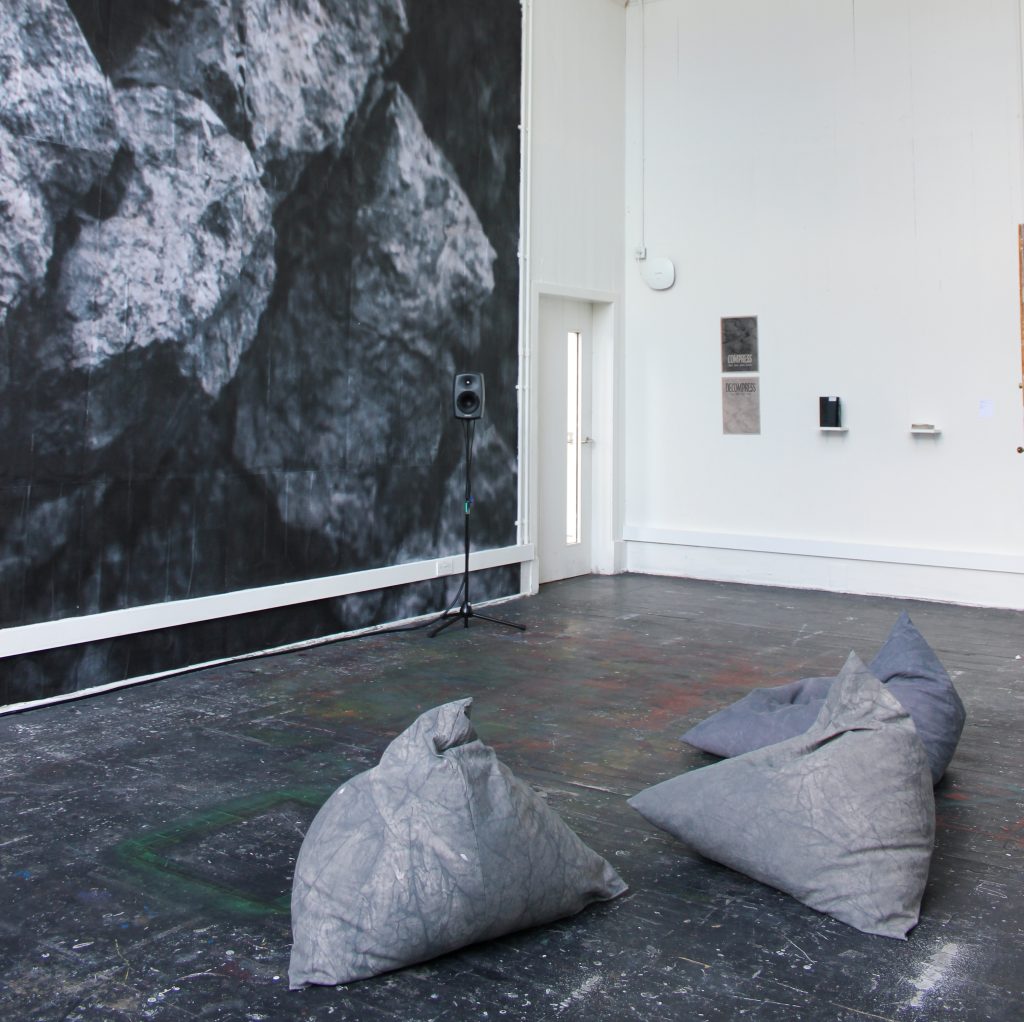 A large room with a wallpapered image of a rock face on one of the walls. Bean bags sit in the middle of the room surrounded by 4 speakers, on the far wall is some supporting print materials about the work.