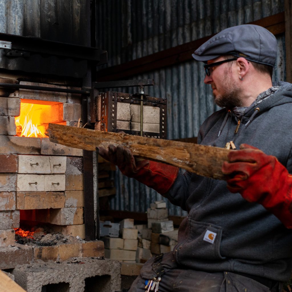A man puts a long piece of wood into a kiln. He is wearing a grey hoody and cap and red gloves.