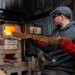 A man puts a long piece of wood into a kiln. He is wearing a grey hoody and cap and red gloves.