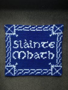 A blue and white crocheted square tapestry that reads “Slàinte Mhath”. The corners of the tapestry each have a Scotland flag. A Celtic braid follows the boarders of the square to connect each flag.
