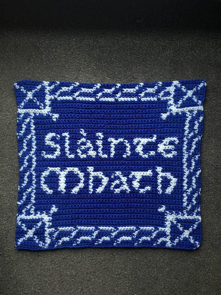 A blue and white crocheted square tapestry that reads “Slàinte Mhath”. The corners of the tapestry each have a Scotland flag. A Celtic braid follows the boarders of the square to connect each flag.