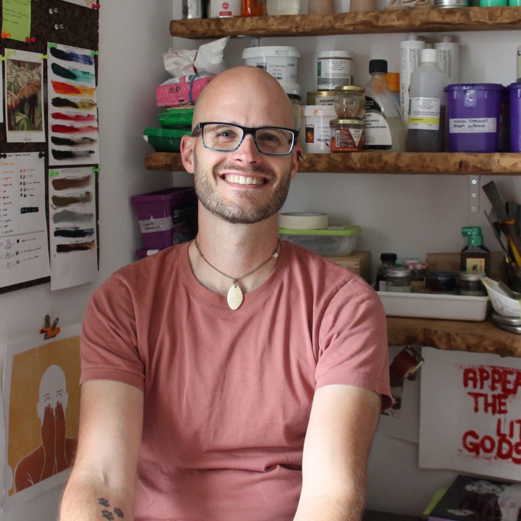 A person smiling in an art studio. They are wearing glasses and a pink t-shirt and there are shelves behind them.