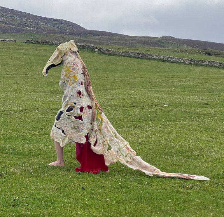A figure with the skull of a horse and a decaying felted body that trails behind it, with one human leg showing, walks across a grassy field with a stone wall in the background.