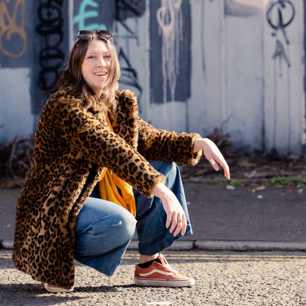 The artist, a young white woman, is crouched outside in front of a wall of graffiti and is wearing a leopard print jacket.