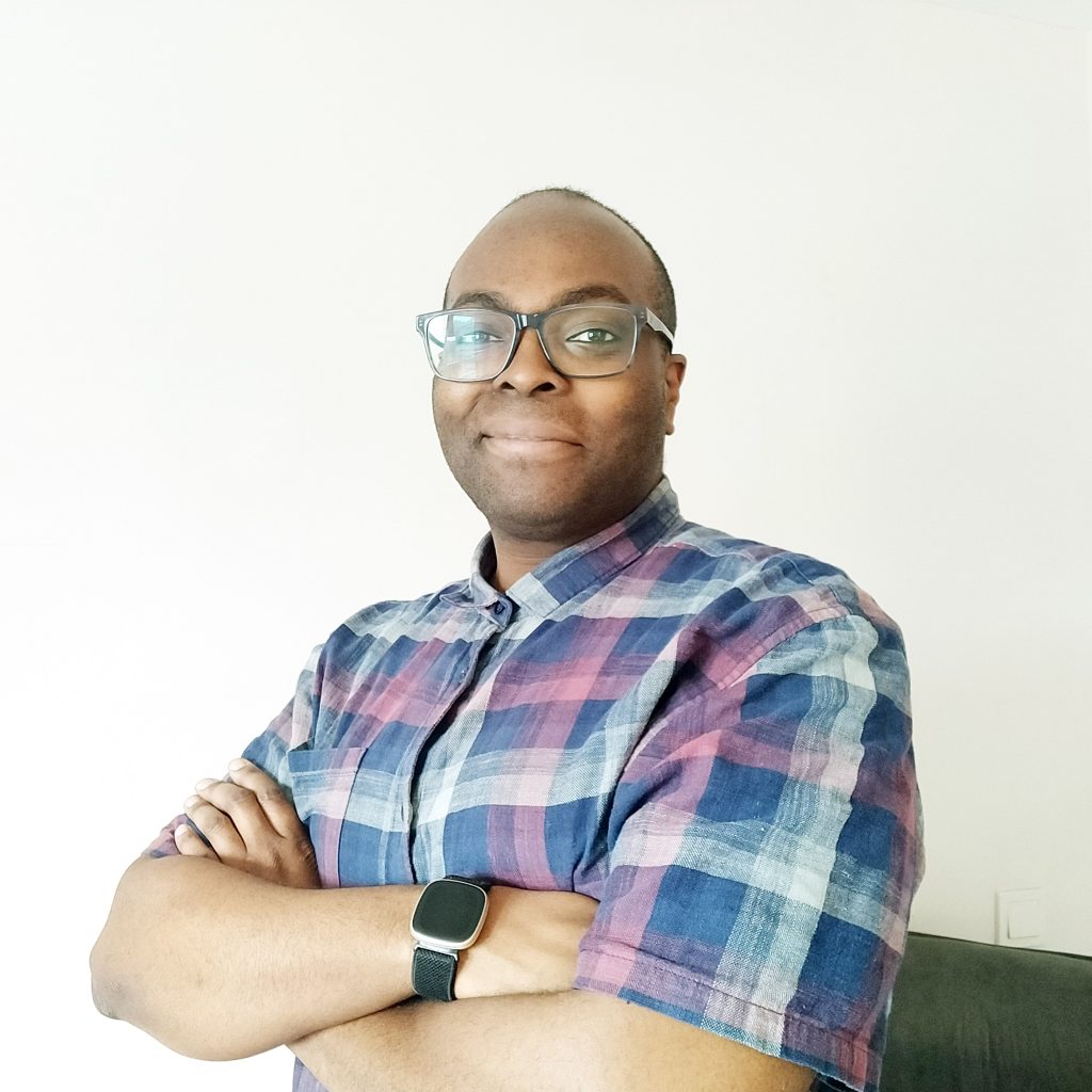 Black man, wearing glasses, smiling, in plaid shirt, against white backdrop.