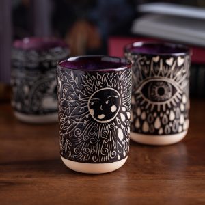 Three handmade ceramic cups sitting on a dark wood surface. The cups are illustrated in black and white with celestial, elemental and nature motifs.