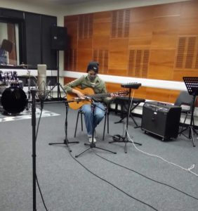 Person sitting in recording studio playing a guitar.