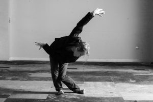 In black and white, a dancer pulls an extended and contorted shape in a sparse indoor space.