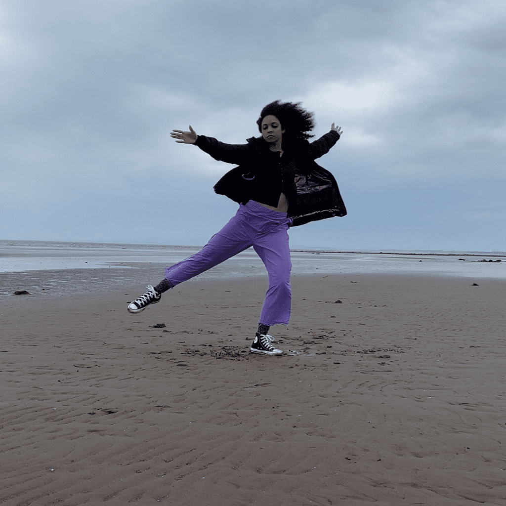 A person dancing on the beach wearing a black jacket and purple trousers.